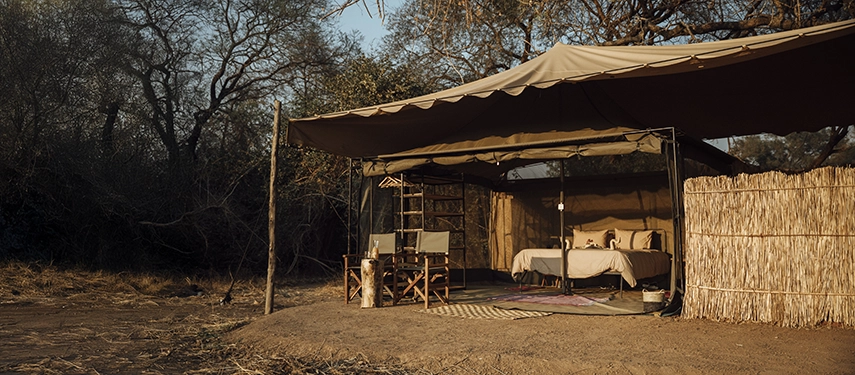An open-fronted safari tent at Kutandala Camp features canvas awnings, outdoor seating and a bedroom immersed in surrounding woodland.