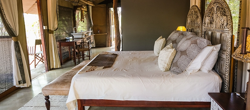 A light-filled safari tent bedroom at Kutandala Camp with carved timber details, open canvas walls and views into the surrounding bush.