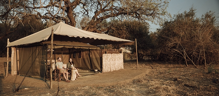 Guests relax on the veranda of a safari tent at Kutandala Camp, enjoying quiet moments framed by mature trees and open bush.