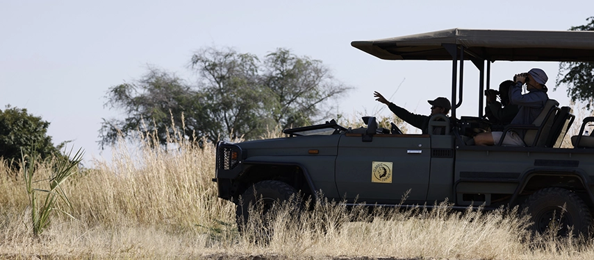 A safari vehicle pauses in tall grass as guides and guests scan the landscape with binoculars during a game drive in North Luangwa.
