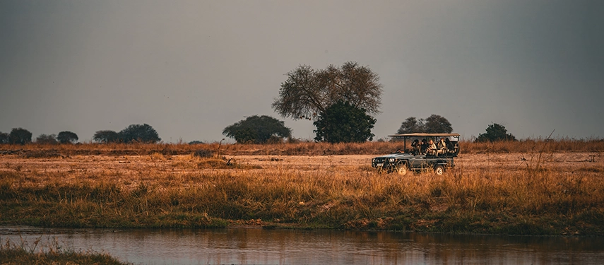 A safari vehicle pauses beside a riverbank in open grassland, offering wide views across North Luangwa’s floodplain habitats.