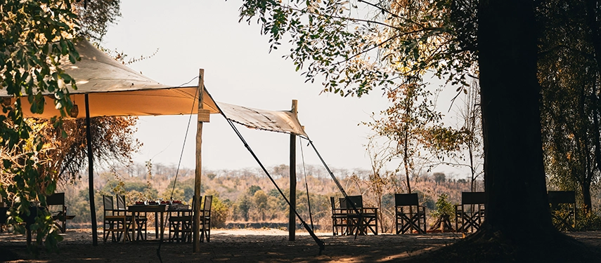 An open-air dining area shaded by canvas and trees is set for meals overlooking the wilderness at Kutandala Camp.