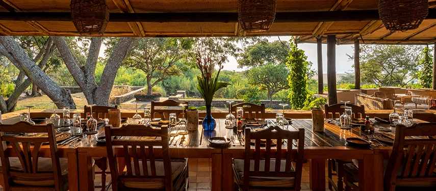 An open-air dining area at Kyambura Gorge Lodge with long communal tables set beneath a shaded veranda overlooking the surrounding bush.