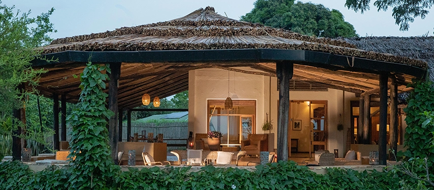 The open-sided main lodge at Kyambura Gorge Lodge glows at dusk, with thatched roofing, warm lighting and views into the surrounding greenery.