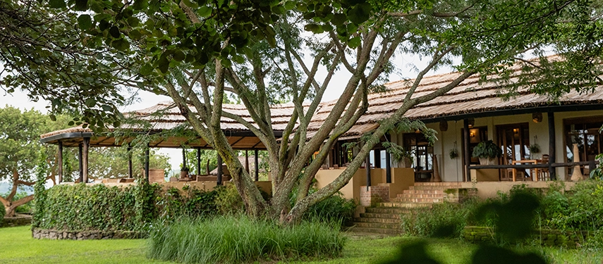 The open-sided main lodge at Kyambura Gorge Lodge sits beneath mature trees, surrounded by gardens and views into the forested landscape.