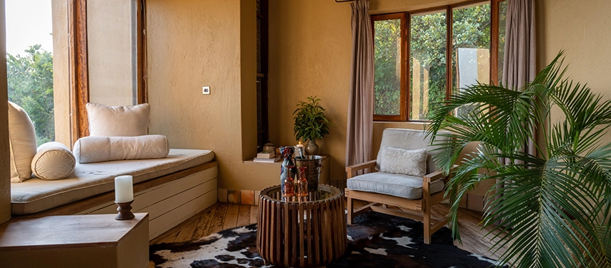 A relaxed indoor sitting area at Kyambura Gorge Lodge featuring soft furnishings, natural textures and leafy forest views through wide windows.