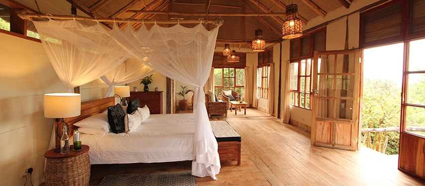 A spacious guest suite at Kyambura Gorge Lodge features a four-poster bed, mosquito netting and natural light flowing in from wide forest-facing doors.