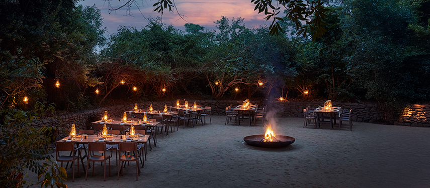Magical open-air boma dining area with firepit and tables set beneath lantern-lit trees.