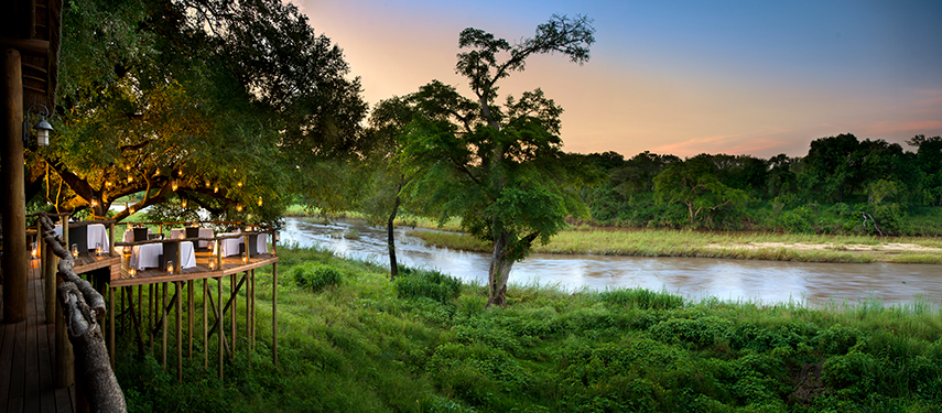 Romantic al fresco dining setup on a lantern-lit deck above the Sabie River at dusk.