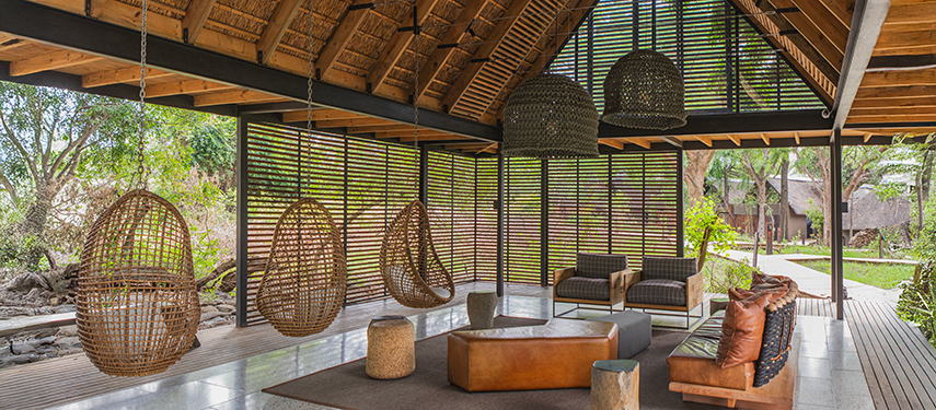 Open-air lounge area with hanging wicker chairs and leather sofas beneath a thatched roof.
