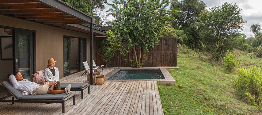 Guests relaxing in a riverside pool surrounded by striped cushions and shaded by umbrellas.