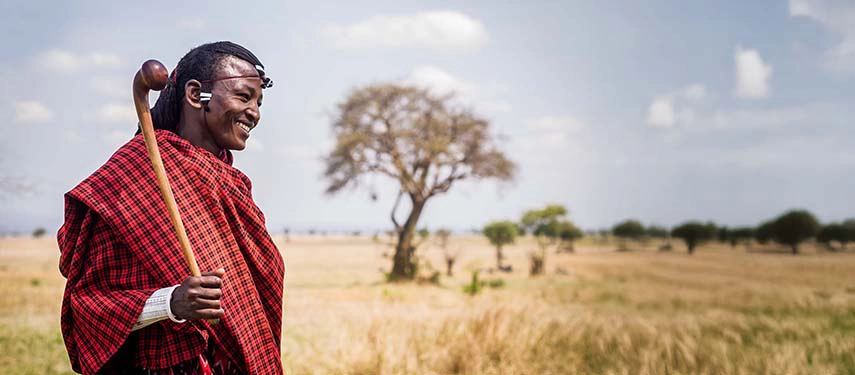 Smiling Maasai man in traditional red shuka standing in the savannah grasslands of Kenya with an acacia tree in the background.