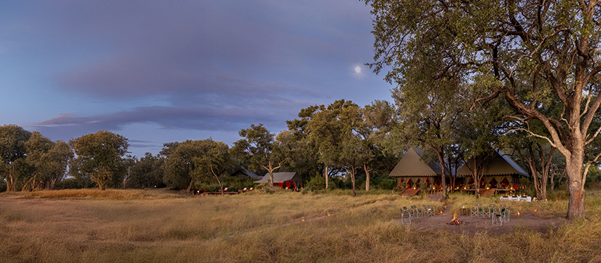 Panoramic view of Mbamba Camp at dusk with firepit and softly lit tents.