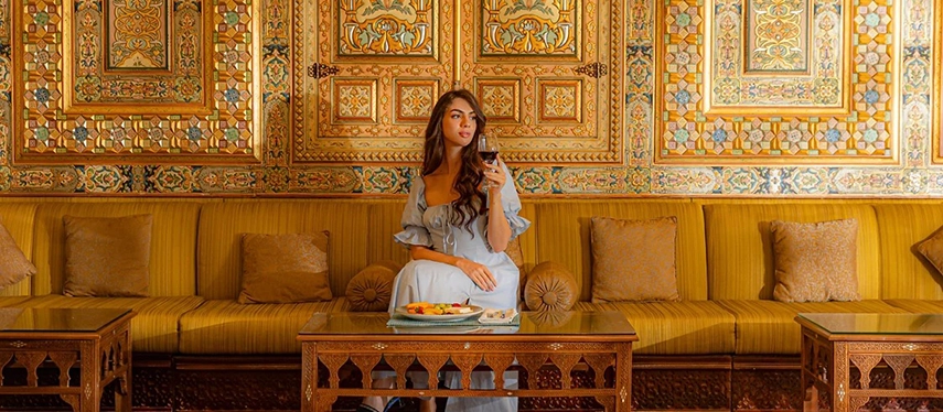 A woman relaxes on a cushioned banquette at Mövenpick Hotel Petra, surrounded by richly detailed mosaic walls and traditional carved wood furnishings.