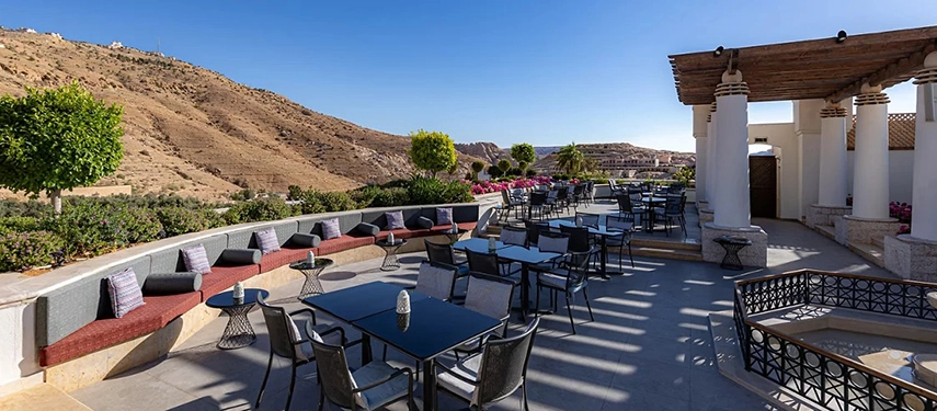 An al fresco terrace at Mövenpick Hotel Petra overlooks the surrounding hills, with shaded seating and tables set beneath clear blue skies.