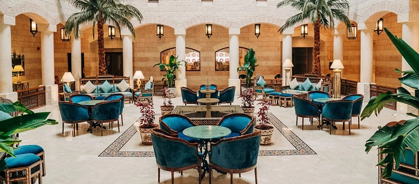 The palm-lined courtyard lounge at Mövenpick Hotel Petra features plush seating arranged around a central fountain within arched stone walls.