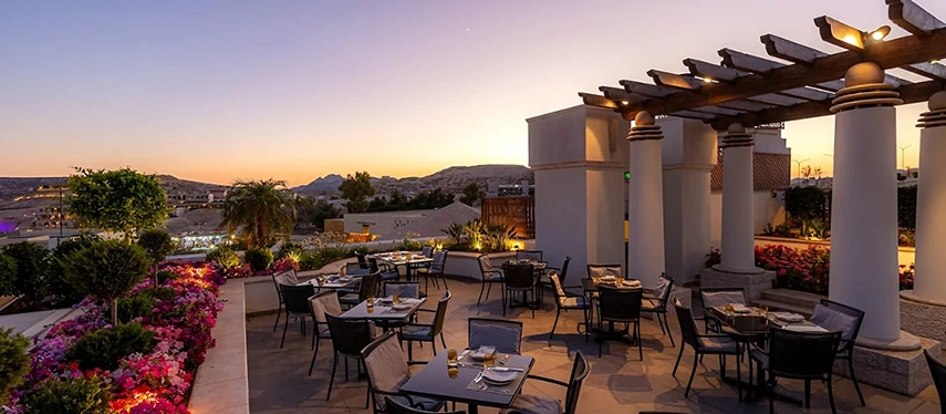 A rooftop dining terrace at Mövenpick Hotel Petra glows at sunset, with tables set among flowering gardens and views across the hills.