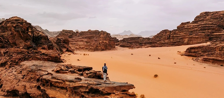 A lone traveller stands on a rocky outcrop in Wadi Rum, gazing across sweeping desert sands and dramatic sandstone formations.