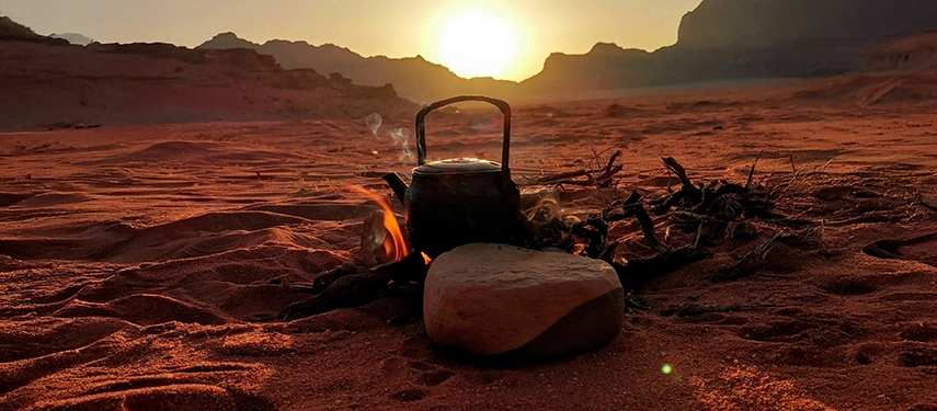 A kettle warms over an open fire in the Wadi Rum desert, silhouetted against glowing sand and distant mountains at sunset.