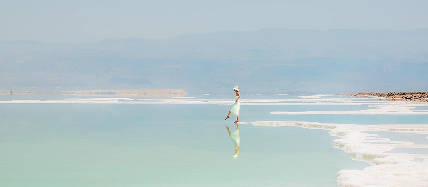 A woman walks across the glassy shallows of the Dead Sea, reflected in pale turquoise waters beneath a hazy sky.