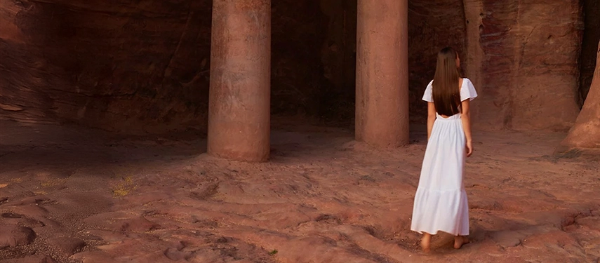 A woman in a flowing white dress walks through Petra’s ancient rock-cut passageway, framed by towering sandstone columns.