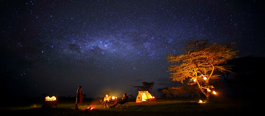 A tourist talks to a guide by a camp fire in Africa at night