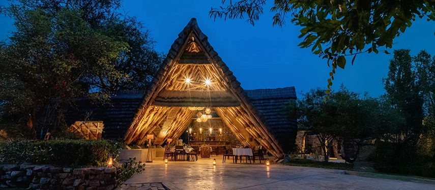 The open-sided dining area glows at dusk, with lantern light illuminating the striking A-frame architecture of the lodge.