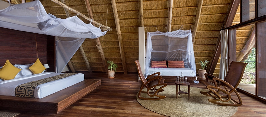 An elegant open-plan suite featuring a raised timber bed, soft lighting and woven textures beneath a high thatched ceiling.