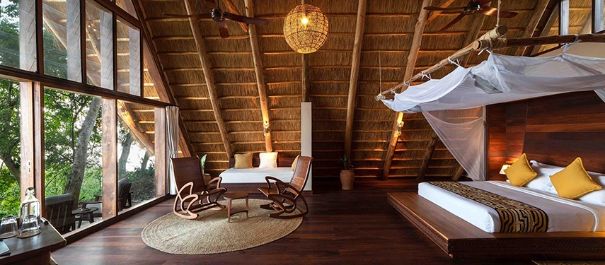A spacious thatched safari suite with timber floors, mosquito-draped bed and lounge seating, opening onto a private deck overlooking riverine forest.