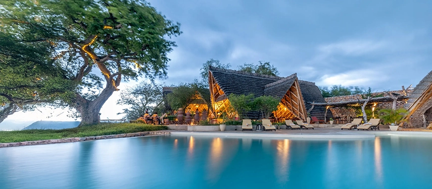 The lodge swimming pool reflects the evening sky, with softly lit thatched buildings and ancient trees creating a tranquil riverfront scene at dusk.