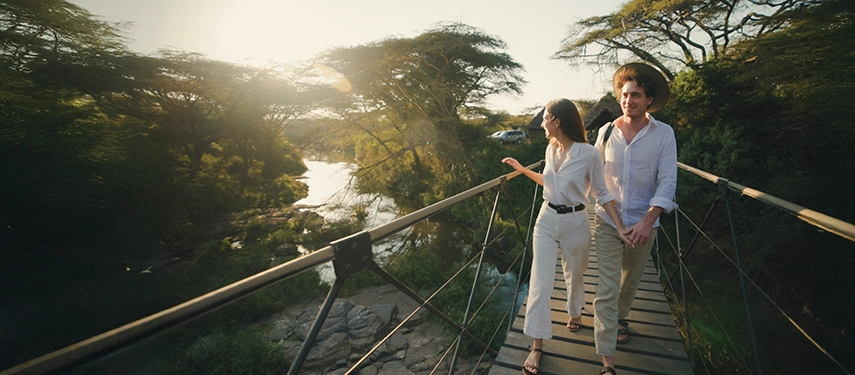 A couple walking hand in hand across Ololo’s footbridge at sunset, surrounded by acacia trees and the gentle flow of the Mbagathi River.