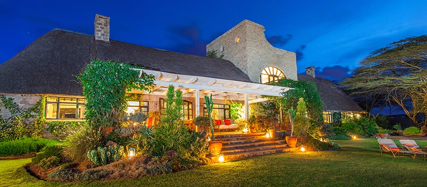 The main house of Ololo Safari Lodge glowing under a deep blue evening sky, surrounded by manicured lawns and softly lit gardens.