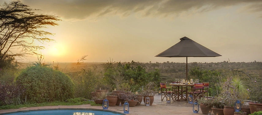 Elegant al fresco dining setup beside the pool at Ololo Safari Lodge, lanterns glowing softly as the sun sets over the plains.
