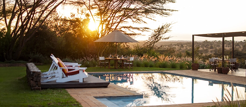 Sun loungers and dining tables arranged by the pool at Ololo Safari Lodge, framed by acacia trees and a glowing evening sky.