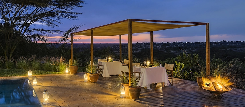 Lantern-lit pool deck at Ololo Safari Lodge set for intimate evening dining, overlooking the darkening savannah under a deep blue sky.