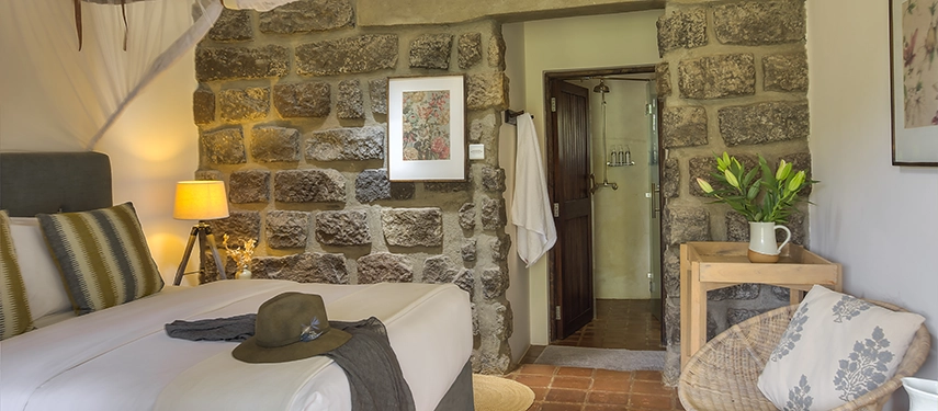 Cosy stone-walled bedroom with soft lighting, earthy tones, and a view into the en suite bathroom at Ololo Safari Lodge’s converted stable wing.