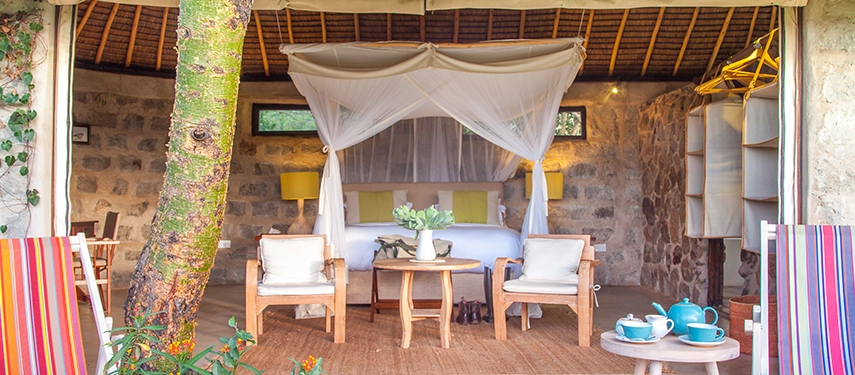 Inviting tented cottage with open front, stone walls, and a canopy bed framed by natural tones and garden views at Ololo Safari Lodge.