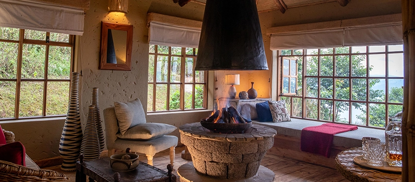 A cosy sitting room at Virunga Lodge features a central fire, deep sofas and panoramic windows framing the green slopes beyond.