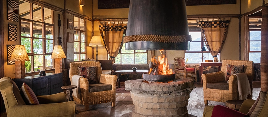 The main lounge at Virunga Lodge centres on a stone fireplace, with woven armchairs, warm lighting and wide windows overlooking the volcanic highlands.