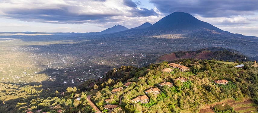A sweeping aerial view places Virunga Lodge on a ridgeline above the Rwandan countryside, with Mount Muhabura dominating the horizon.
