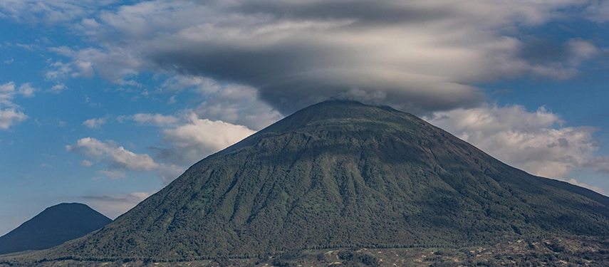 Mount Muhabura rises dramatically above the landscape near Virunga Lodge, its volcanic slopes crowned with cloud against the blue Rwandan sky.