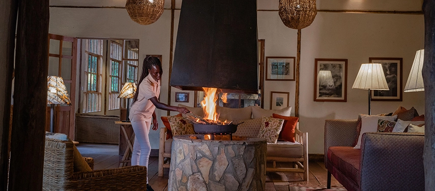 The main lounge at Volcanoes Bwindi Lodge, where a staff member tends a central fireplace beneath woven lanterns, creating a warm and welcoming atmosphere.