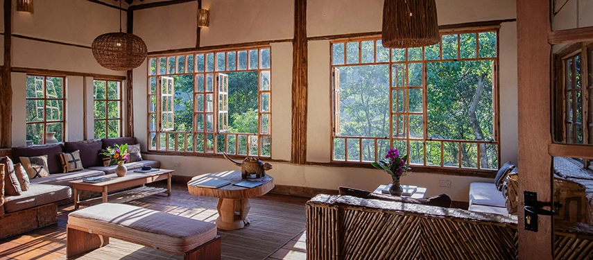 A bright communal lounge at Volcanoes Bwindi Lodge with expansive windows, handcrafted furniture and tranquil views into the surrounding rainforest.