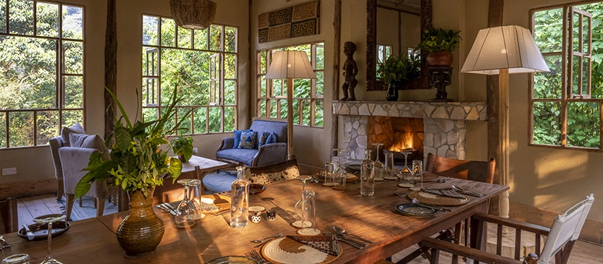A warmly lit dining room at Volcanoes Bwindi Lodge with a long timber table, stone fireplace and forest views through tall windows.