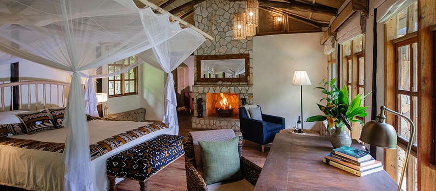 A spacious suite interior at Volcanoes Bwindi Lodge, featuring a four-poster bed, stone fireplace and forest views through wide timber-framed windows.