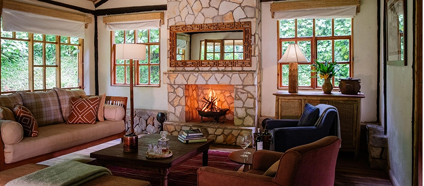 The main lounge at Volcanoes Bwindi Lodge, with a crackling fireplace, comfortable seating and large windows overlooking the surrounding rainforest.