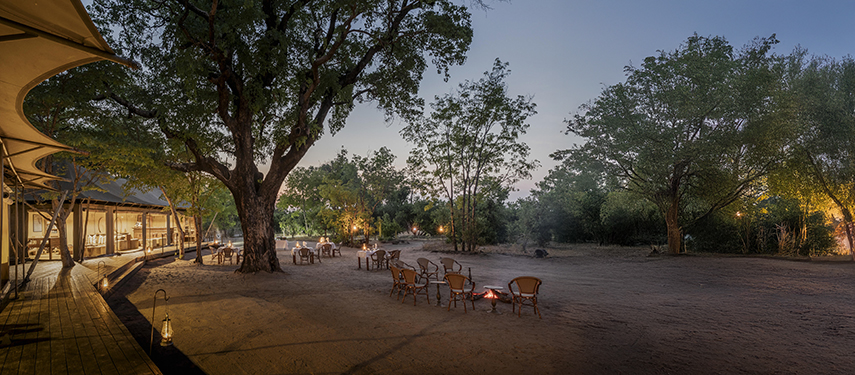 Al fresco dining area under lantern-lit trees at dusk in the main camp clearing.
