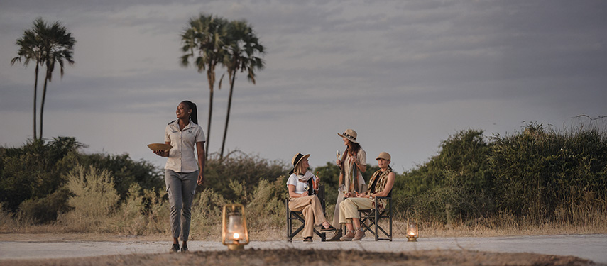 Guests enjoy sundowners near a campfire, with a waitress serving drinks in Botswana's Okavango Delta