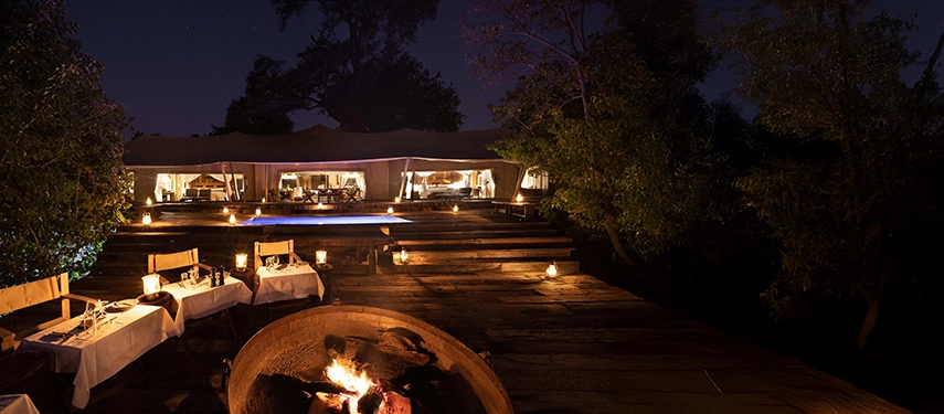 Night-time view of the Zarafa Dhow Suite in the Selinda Reserve, with lantern-lit decks, firepit dining and the tented suite glowing softly under the stars.