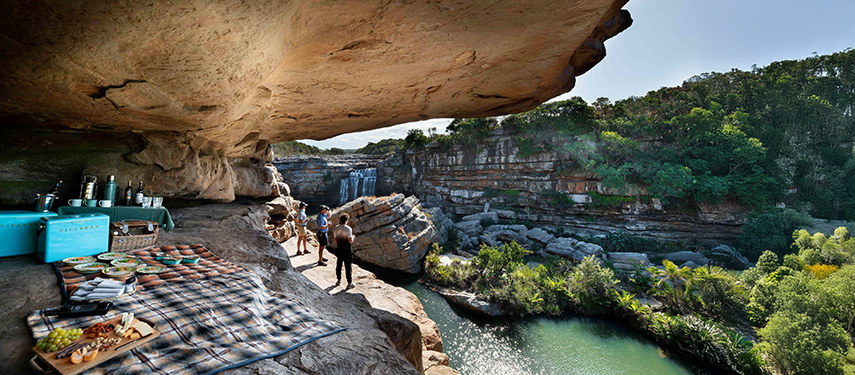 Scenic picnic setup beneath a rock overhang beside a river and waterfall.